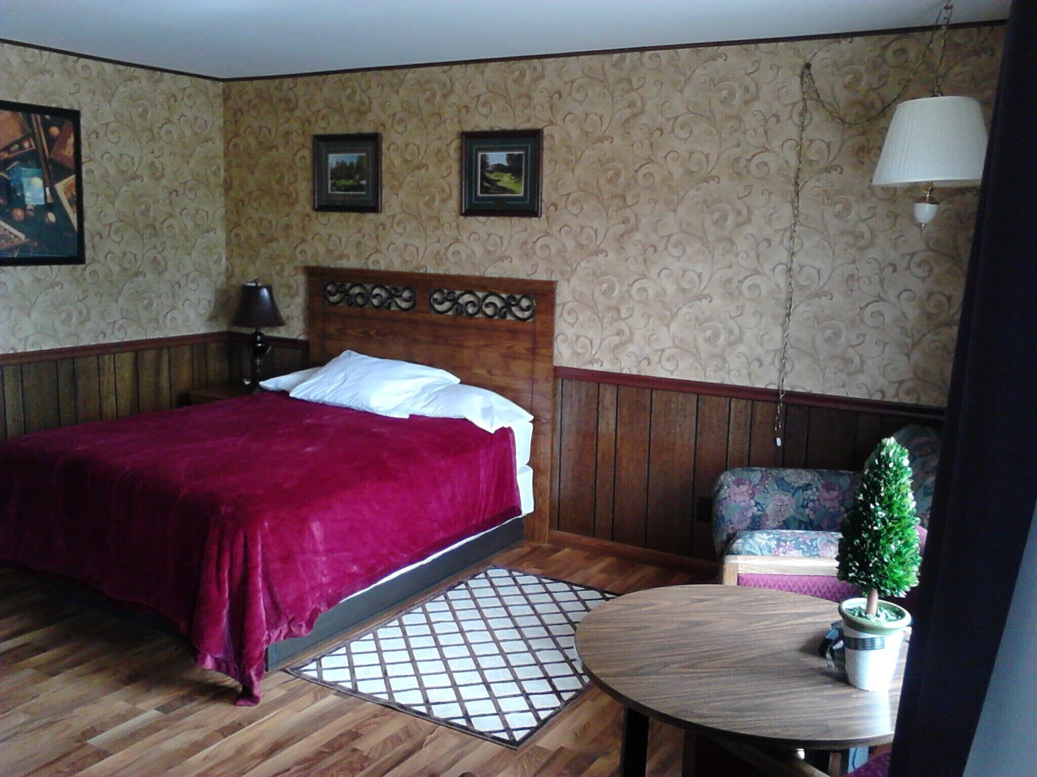 A bedroom with a wooden bed, a small round table with a potted plant, a chair, and a decorative headboard.