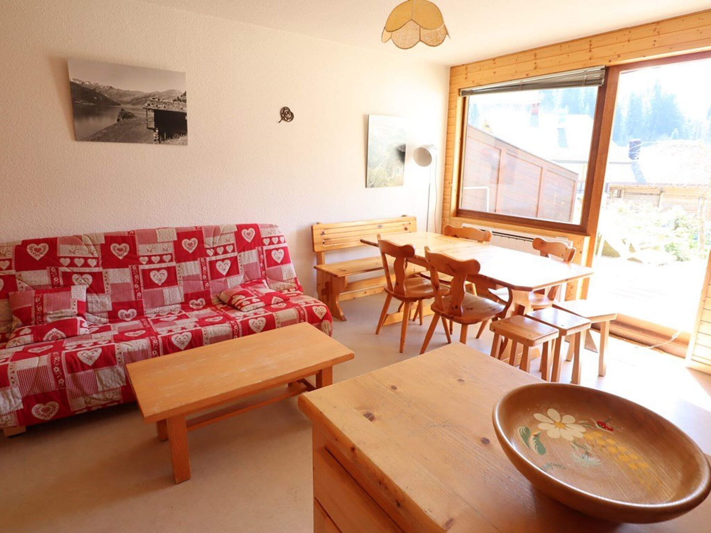 A cozy living room with a red and white patterned sofa, wooden furniture, a dining table, and a large window offering a view of the outdoors.