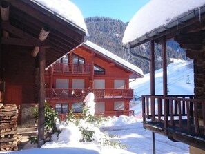 Un chalet de madera con balcón, rodeado de nieve y una pista de esquí al fondo.