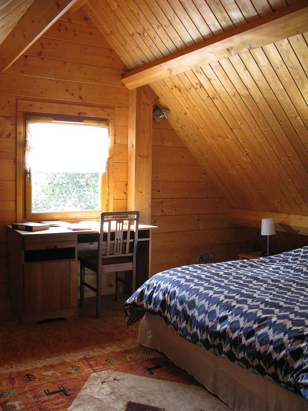 Une chambre mansardée chaleureuse, avec un bureau et une chaise en bois, un lit avec une literie à motifs et une fenêtre laissant entrer la lumière naturelle.