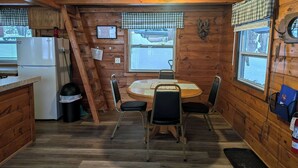 Interior de una cabaña de madera con una mesa redonda, sillas, un refrigerador, una ventana con vistas y una escalera.
