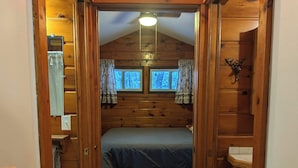 Habitación de cabaña de madera con una cama, dos ventanas con cortinas, una lámpara en el techo y un estante montado en la pared con una decoración de cabeza de venado.