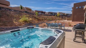 A hot tub with a patio area, outdoor seating, and a view of a hillside with buildings. - Sleeps 16 - Private Hot Tub, Fire Pit & Poolside (St. George)