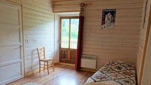 A cozy cabin room with a wooden floor, a bed with a quilt, a chair, and a door leading outside.