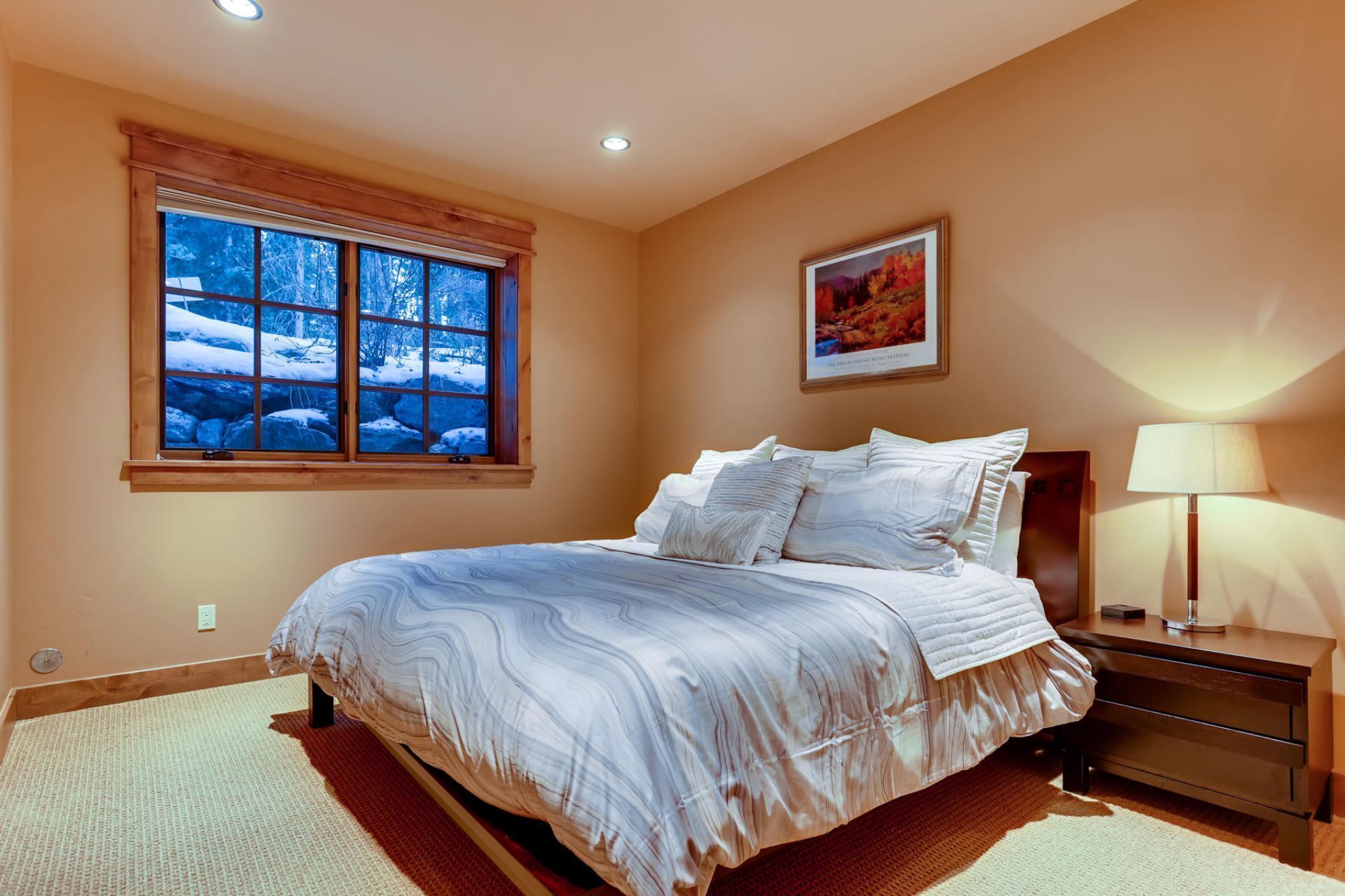A bedroom with a large window, a bed, a nightstand, and a painting on the wall.