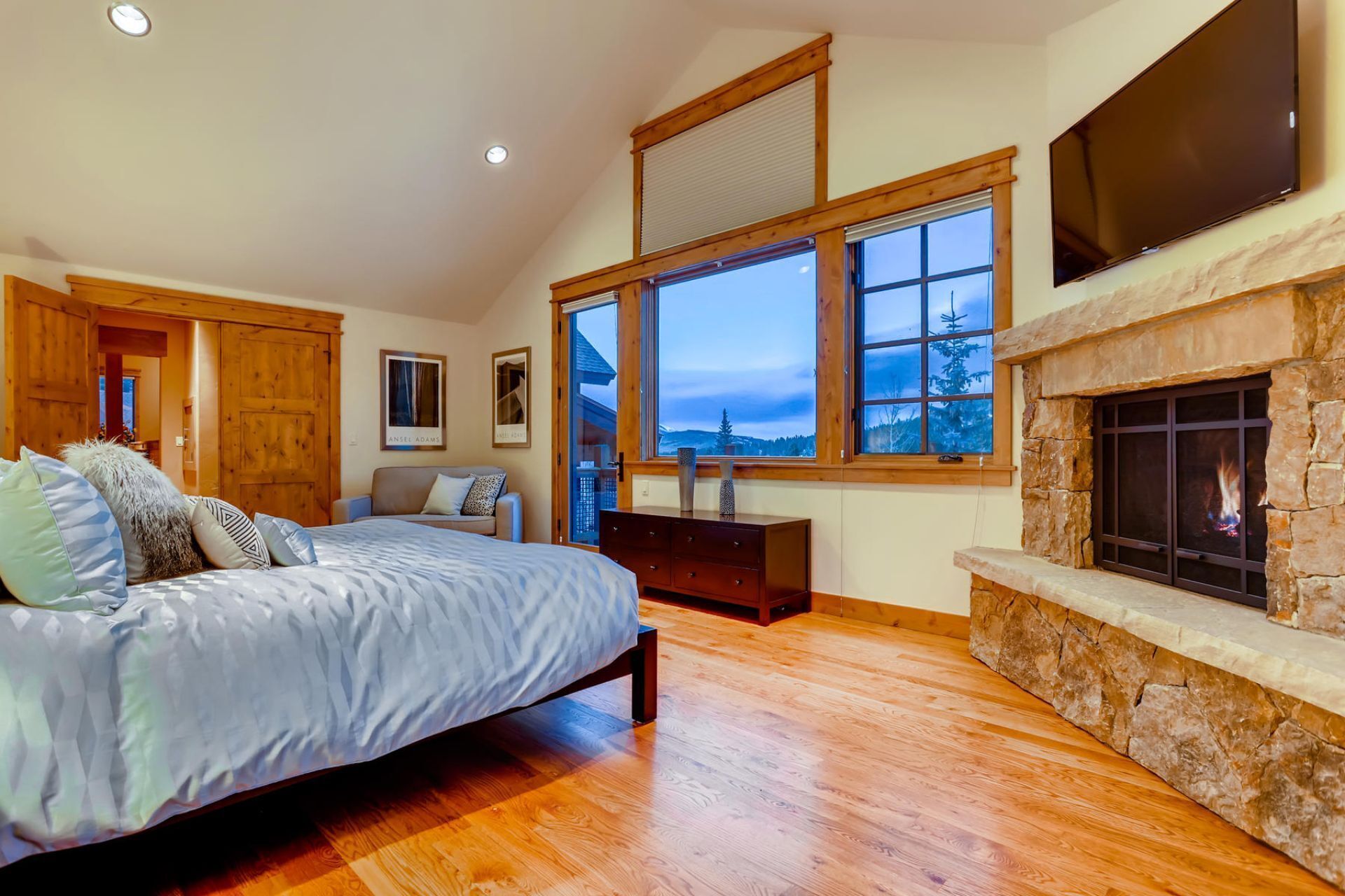 A bedroom with a bed, a dresser, a fireplace, and large windows with a view of trees and mountains.