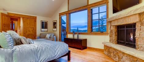A bedroom with a bed, a dresser, a fireplace, and large windows with a view of trees and mountains.