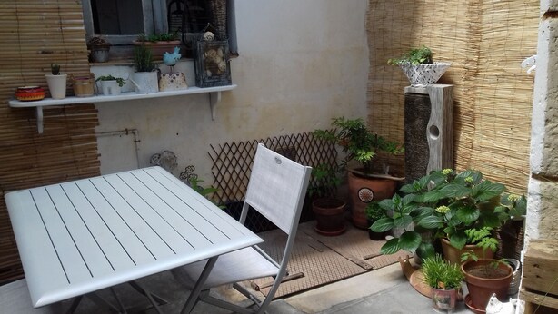 Un petit patio extérieur avec une table et une chaise en métal, entouré de plantes en pot et agrémenté d’un mur décoratif.