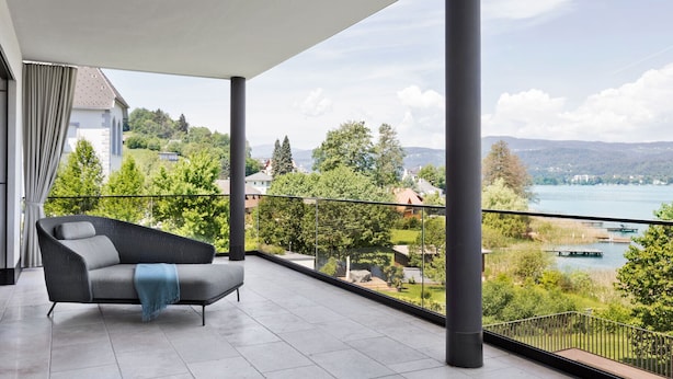 A balcony with a grey sofa and a view of a lake and surrounding greenery.