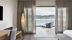 A hotel room with a bed, a desk, a chair, a floor lamp, and a view of a lake with mountains in the background.