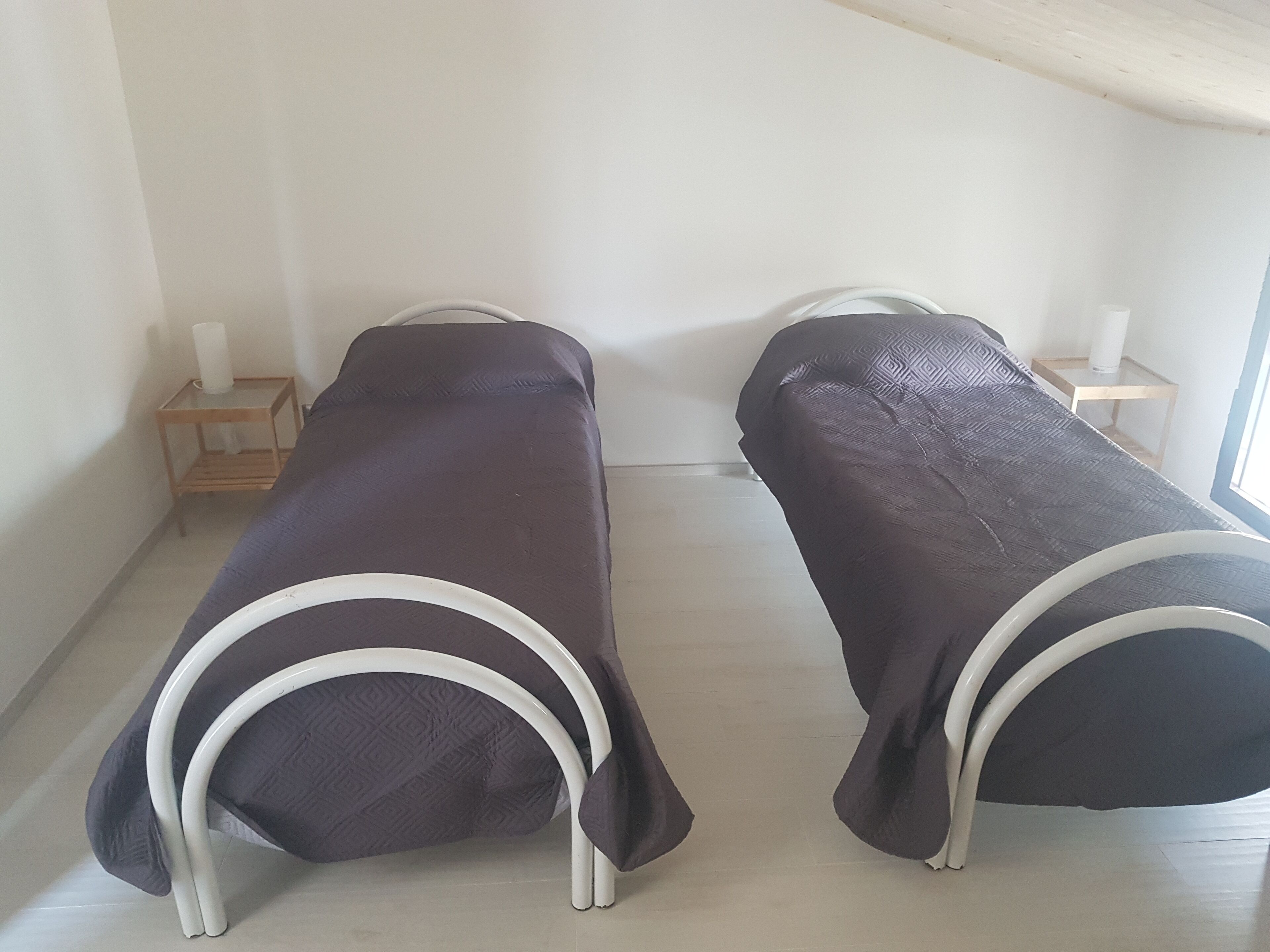 Two single beds with white metal frames and dark covers in a room with white walls and a wooden floor.