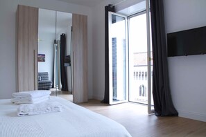 A modern bedroom with a large bed, a wardrobe, a sliding glass door to the balcony, and a flat-screen TV mounted on the wall.