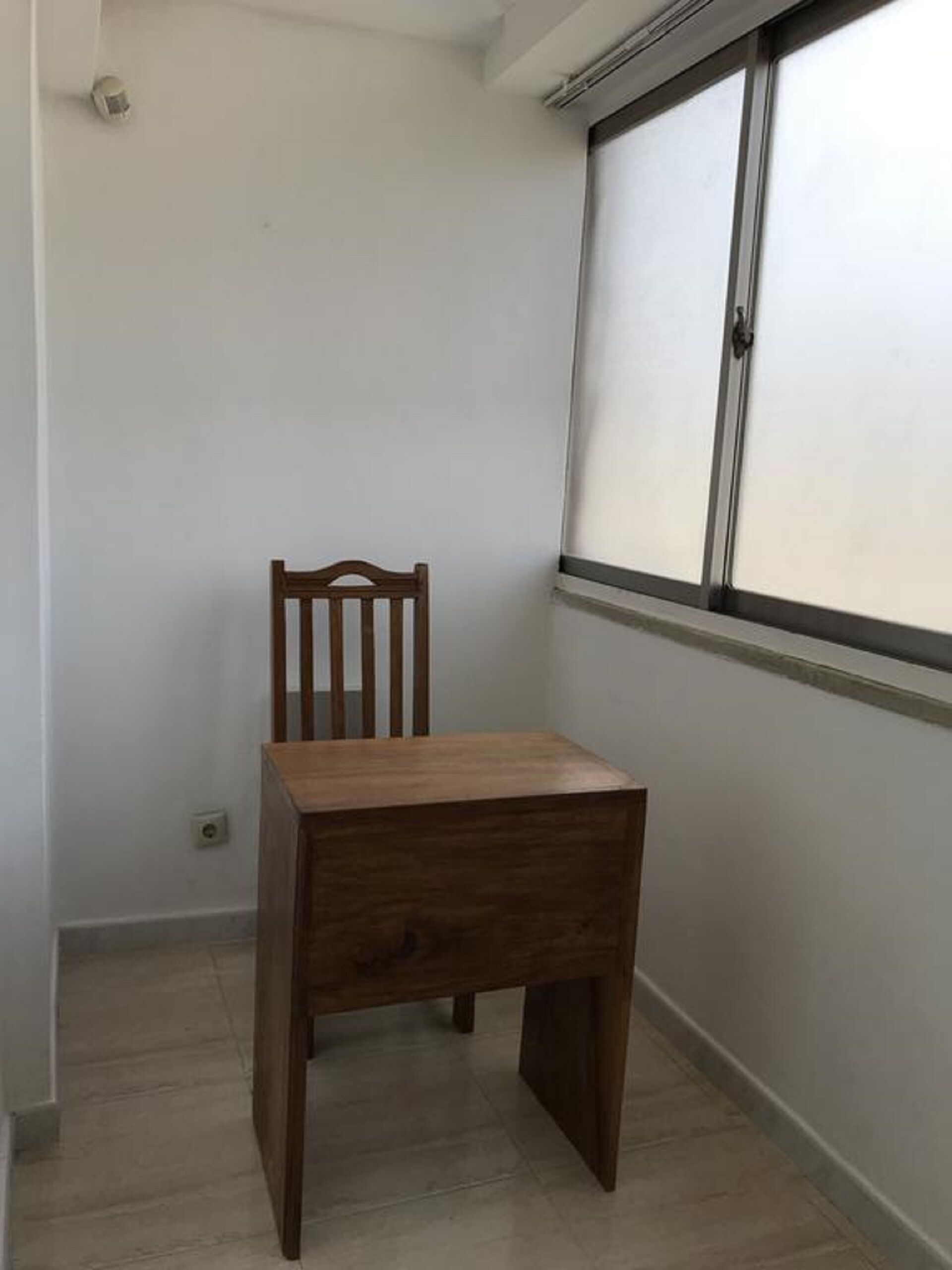A corner room with a wooden chair and a small table, a window with blinds, and a light switch on the wall.
