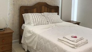 Un dormitori amb un panell de fusta al capçal del llit, un llençol blanc, una tauleta de nit amb una làmpada i un aire acondicionat fixat a la paret.