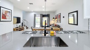 A modern kitchen with a stainless steel sink, a dining area with a table and chairs, a TV mounted on the wall, and a staircase leading to another level. - A Dream Villa next to Disney. (Kissimmee)
