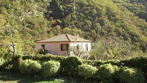 A small, single-story house with a tiled roof, surrounded by dense greenery and hills in the background.