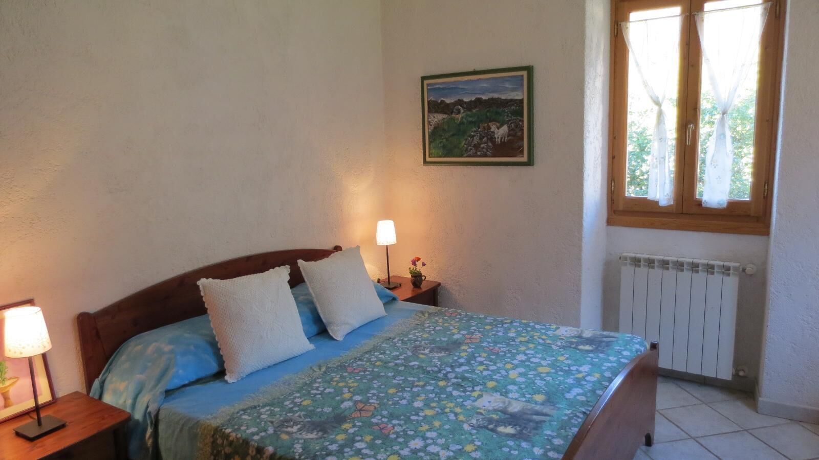 A bedroom with a bed, two bedside lamps, a radiator, and a window with curtains.