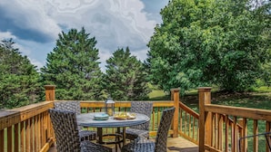 Een houten terras met een ronde tafel en vier stoelen, omgeven door een houten balustrade, met uitzicht op een gazon met bomen.