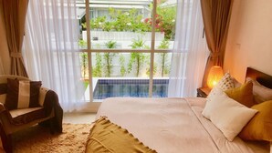 Ein Schlafzimmer mit einem großen Bett, einem Sessel, einem Nachttisch mit Lampe und Blick auf ein Schwimmbad und Grünflächen.