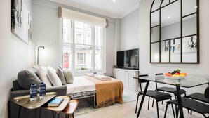 A modern hotel room with a bed, a desk with a laptop, a TV, and a dining area with a fruit bowl. - Comfy Bayswater Apartment (London)