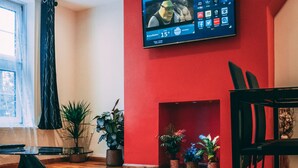 Ein modernes Wohnzimmer mit roter Akzentwand, einem flachbildfernseher über einem Kamin, Holzböden und mehreren Topfpflanzen.
