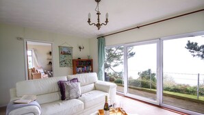 A spacious living room with a white sofa, a wooden coffee table with a bottle and glasses, a bookshelf, and a large window offering a view of greenery. - Windwhistle - Two Bedroom Cottage, Sleeps 4 (Saint Martin-by-Looe)