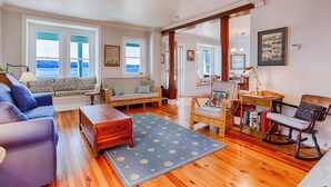 A spacious living room with a large window overlooking a body of water, a wooden floor, and a variety of seating options including a sofa, armchairs, and a rocking chair.