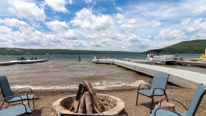 A lakeside dock with chairs and a fire pit, boats on the water, and a dock extending into the lake.