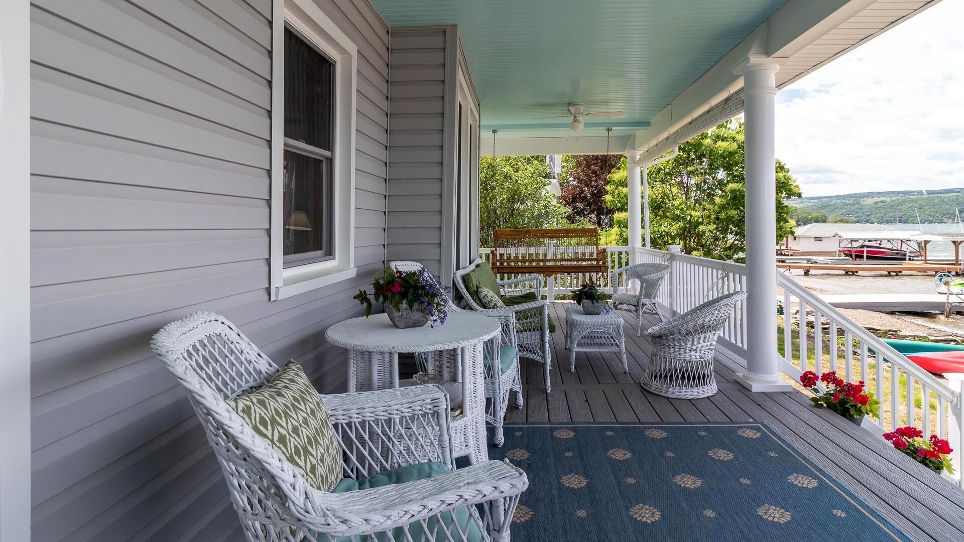 A covered porch with wicker furniture, a small table, and a view of the water and boats in the background.