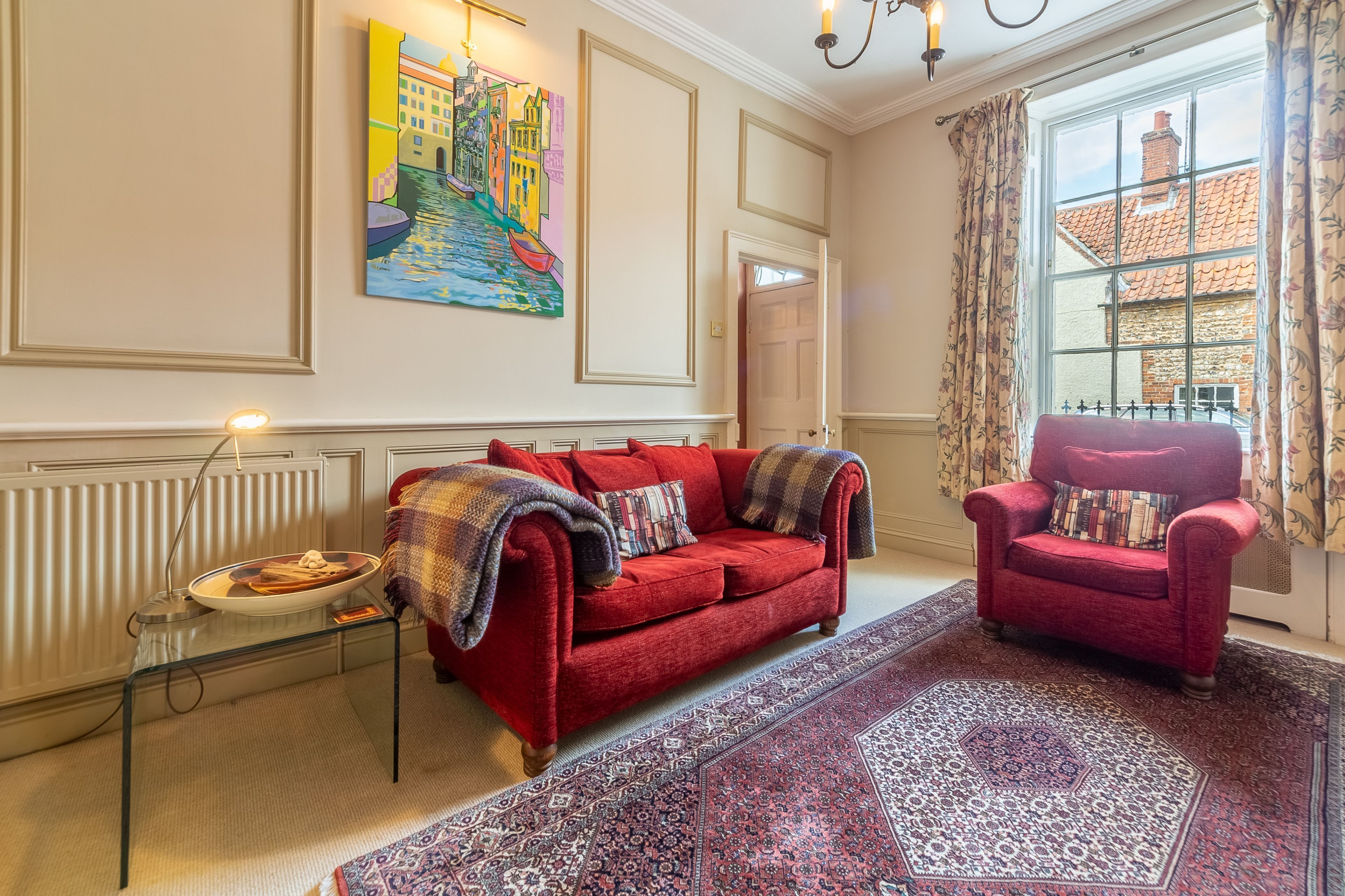 A living room with a red sofa, a red armchair, a large window with a view of buildings, a painting on the wall, and a radiator.