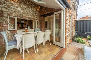 A patio with a stone wall, a dining table with chairs, and a sliding glass door leading to a garden. - Estcourt House, Burnham Market, Norfolk (Burnham Market)
