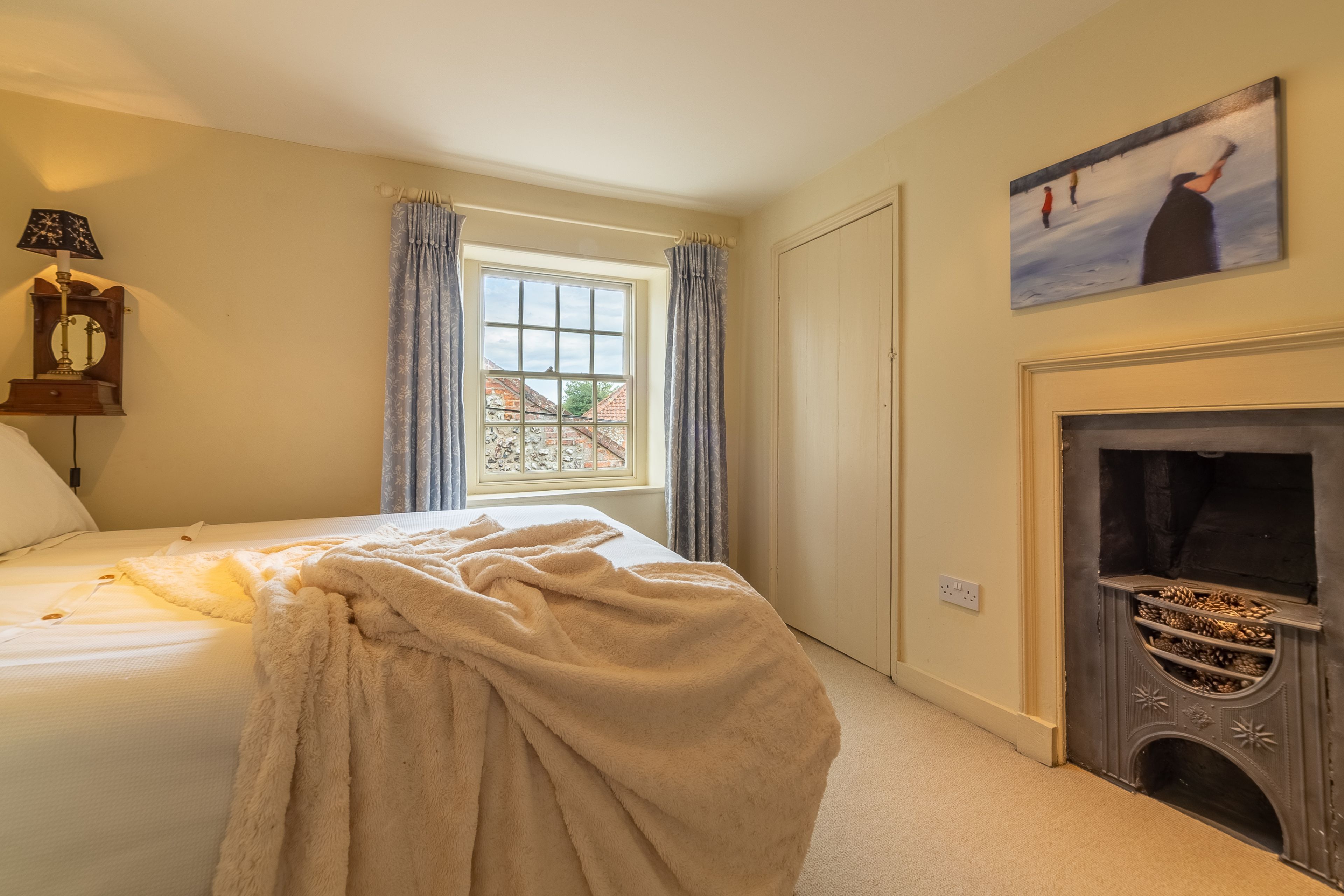 A bedroom with a bed, a window with curtains, a fireplace with pinecones, and a painting on the wall.