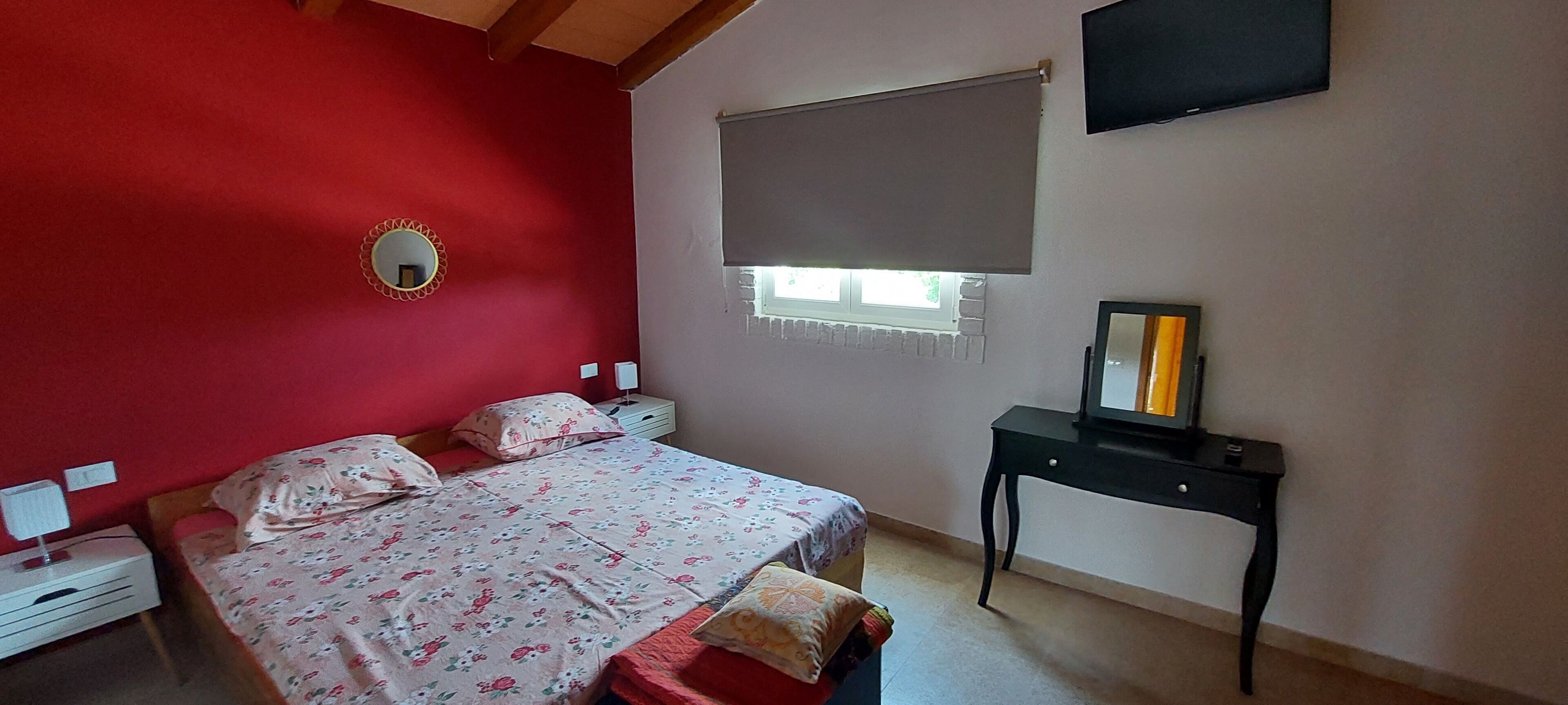 Ein Schlafzimmer mit Bett, Nachttisch, Schreibtisch, Spiegel und Fenster mit Jalousie.