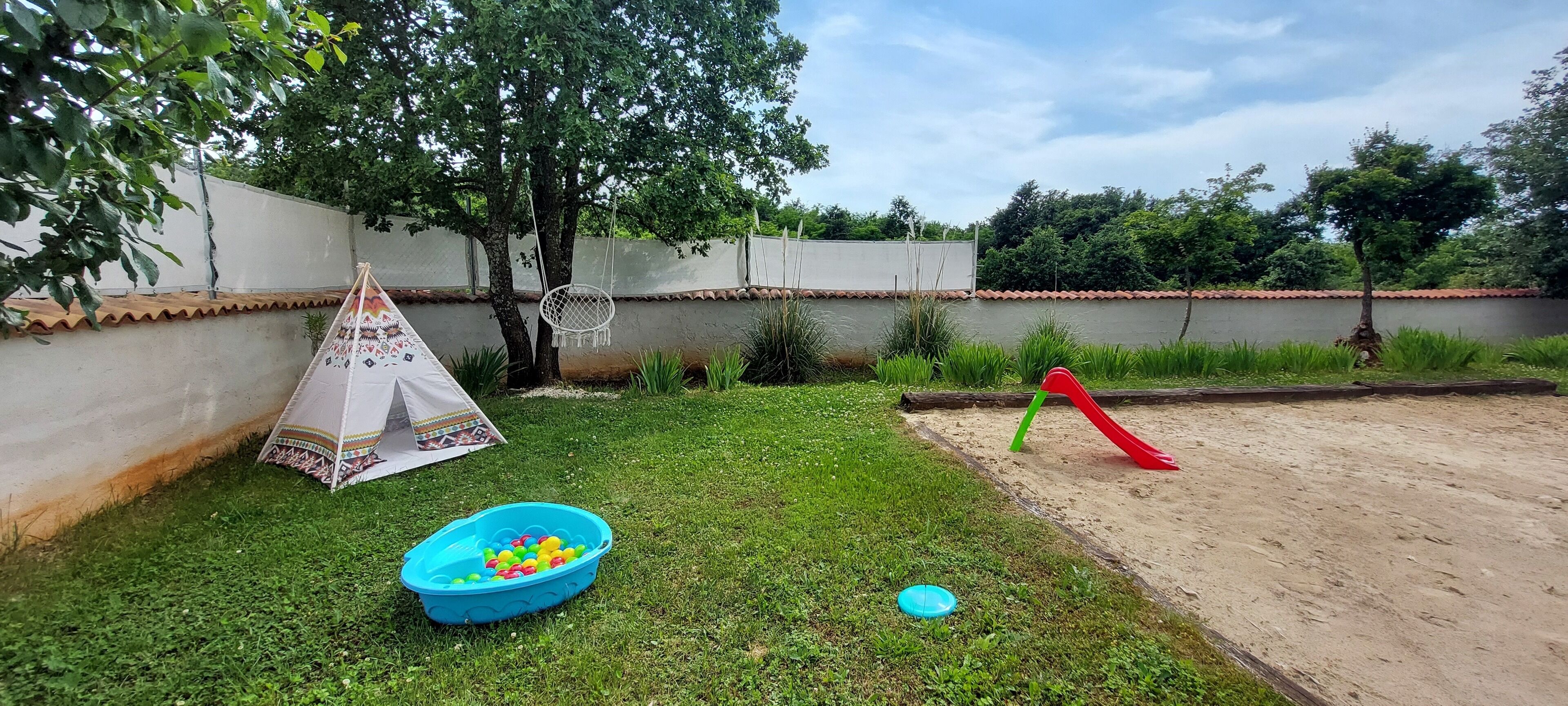 Un área con césped, una carpa de juegos, una piscina de bolas azul y un tobogán rojo.