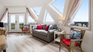 Ein gemütlicher Raum mit grauem Sofa, roten Kissen, Korbstühlen und Blick durch große Fenster.