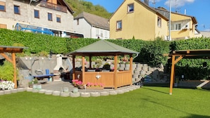 A wooden gazebo with a green roof surrounded by a stone wall, a barbecue grill, and potted plants.