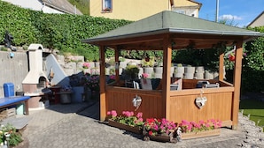 A wooden gazebo with a tiled roof, surrounded by flower beds and a paved patio.
