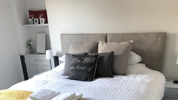 Una cama bien hecha con ropa blanca, un edredón color mostaza y una almohada con una frase motivacional.