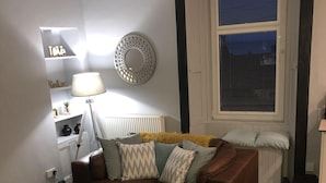 Una sala de estar con un sofá de cuero marrón, una mesa de centro de madera, una planta en maceta, un espejo decorativo y una ventana con persianas.