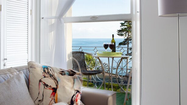 Ein Balkon mit Meerblick, ein Tisch mit zwei Weingläsern und ein Korbstuhl.