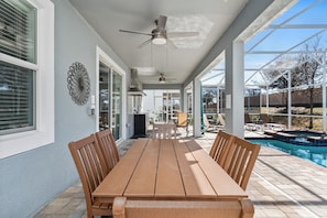 Une terrasse couverte avec une table et des chaises en bois, un ventilateur de plafond et une vue sur une piscine.