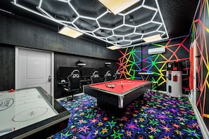 Une pièce équipée d’une table de billard, d’un baby-foot et d’une table de jeu de flipper. On y trouve quatre chaises et un téléviseur. Les murs sont décorés de motifs géométriques colorés.