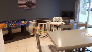 A dining area with a table, chairs, a high chair, a playpen with toys, and a foosball table.