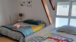 A bedroom with two beds, a wooden ceiling beam, a wall decal, a small table with a lamp, and a window with blinds.