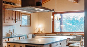 A cozy kitchen with wooden cabinets, a large window with a view of snow-capped mountains, and a hanging light fixture.