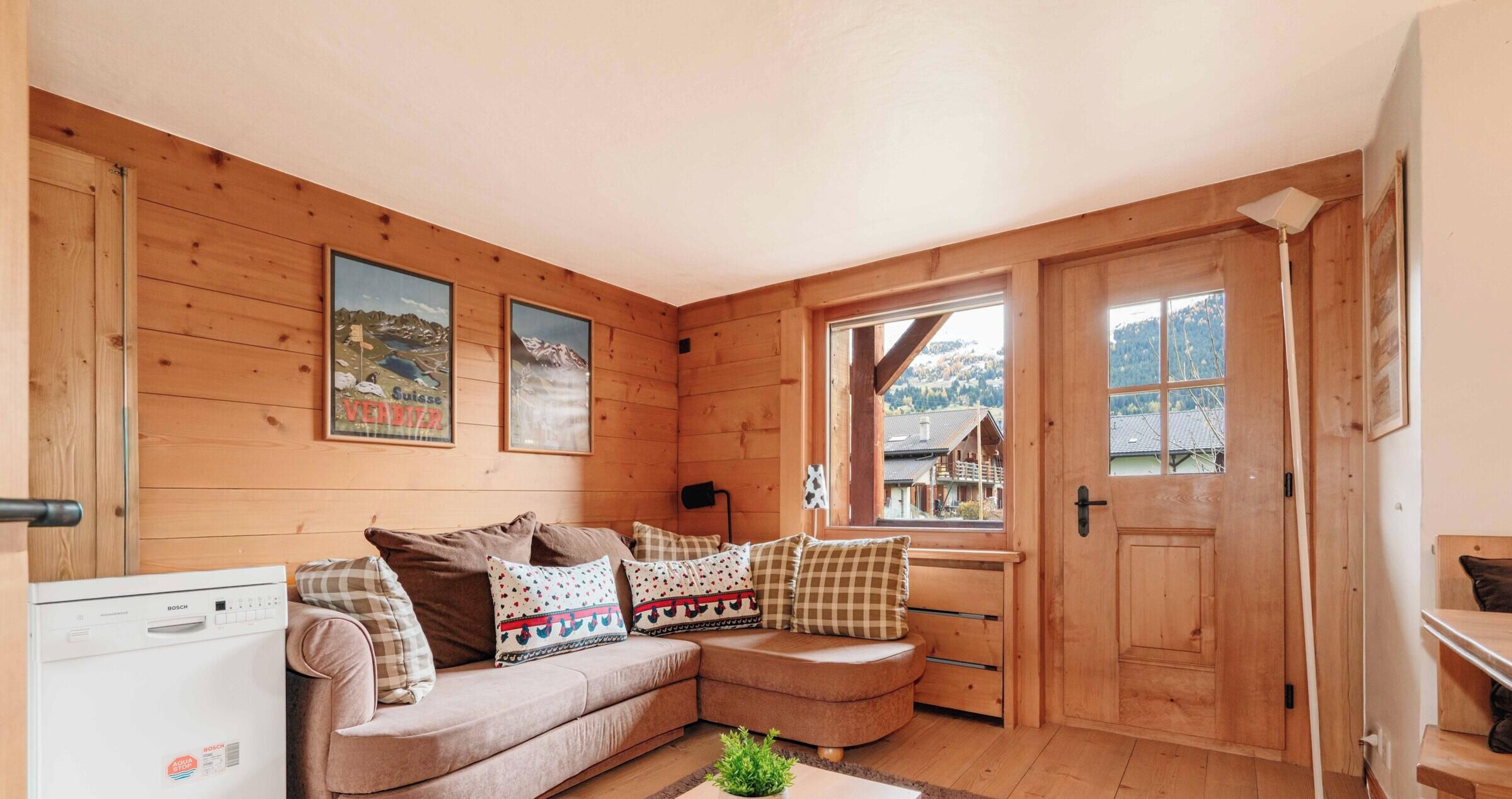A cozy cabin interior with a wooden interior, a sofa with patterned cushions, a Bosch washing machine, and a window offering a view of a mountain landscape.