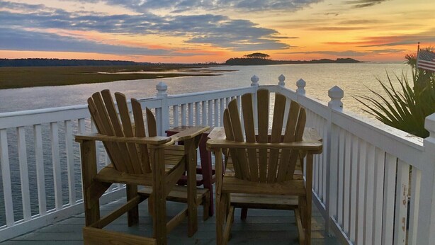 Dos sillones Adirondack de madera sobre una terraza blanca con barandilla, con vista a un cuerpo de agua y una masa de tierra distante al atardecer.