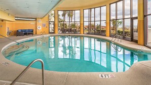 An indoor swimming pool with clear blue water, surrounded by large windows offering views of palm trees and a cloudy sky. - Two Bedroom Ocean View Luxury, Myrtle Beach 1751742 (North Myrtle Beach)