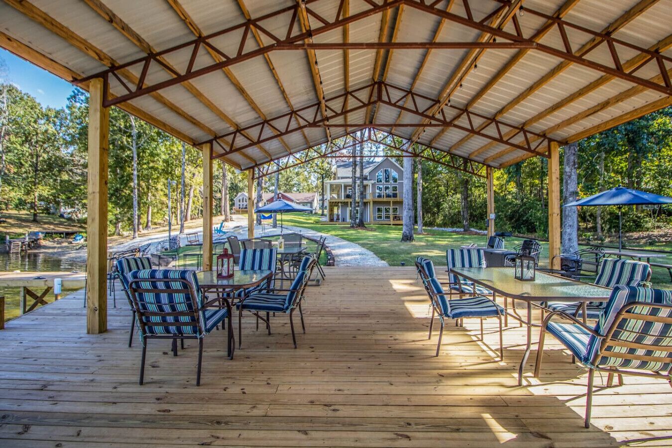A covered outdoor deck with wooden flooring and metal beams, surrounded by trees and a lake.
