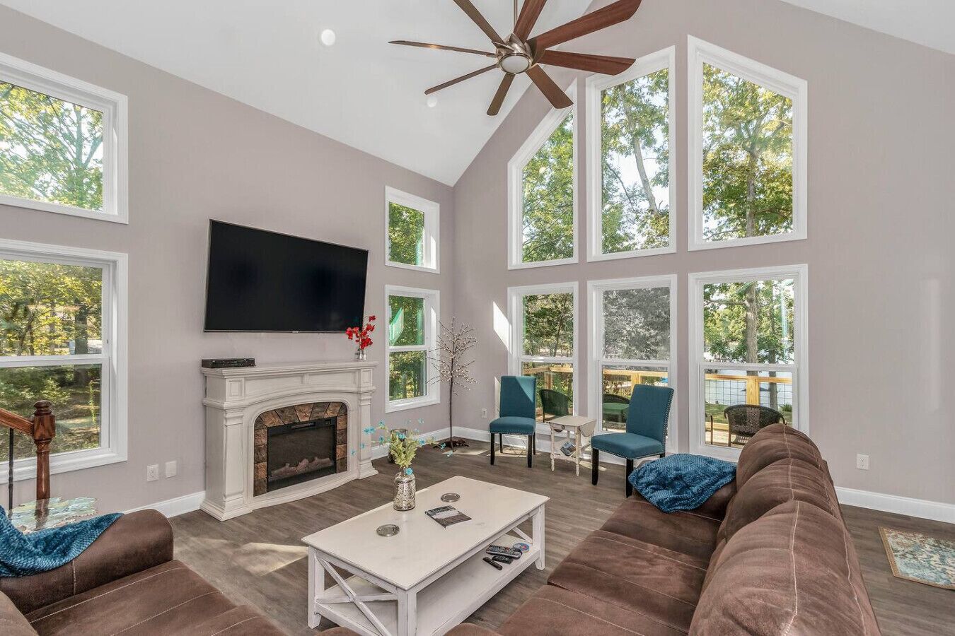 A spacious living room with a fireplace, large windows, a ceiling fan, and a flat-screen TV.
